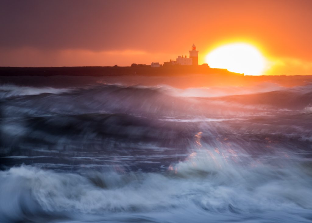 Sunrise behind an island. The rough sea splashes and golden sunlight lights up the spray. 