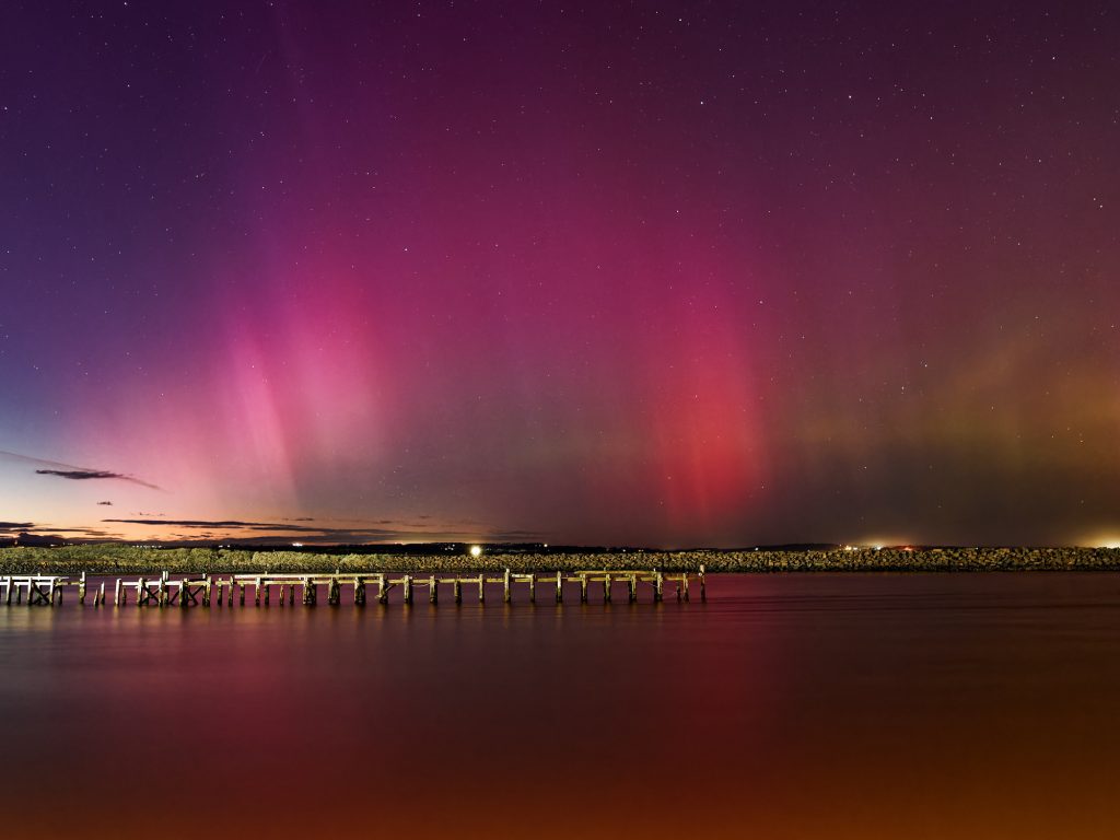 The aurora above a pier is reflected in the sea. Photographed on a nighttime workshop