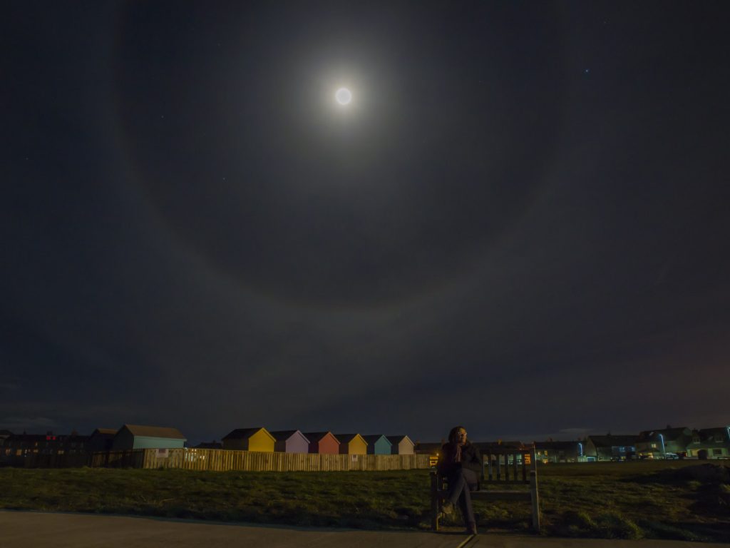 Nighttime shot of a moon halo with colouration, making it a moobow, in the sky above beach huts and my lovely wife sitting on a bench