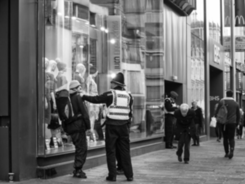 A blurred image of a man under arrest as another man, bent double with age, walks past. 