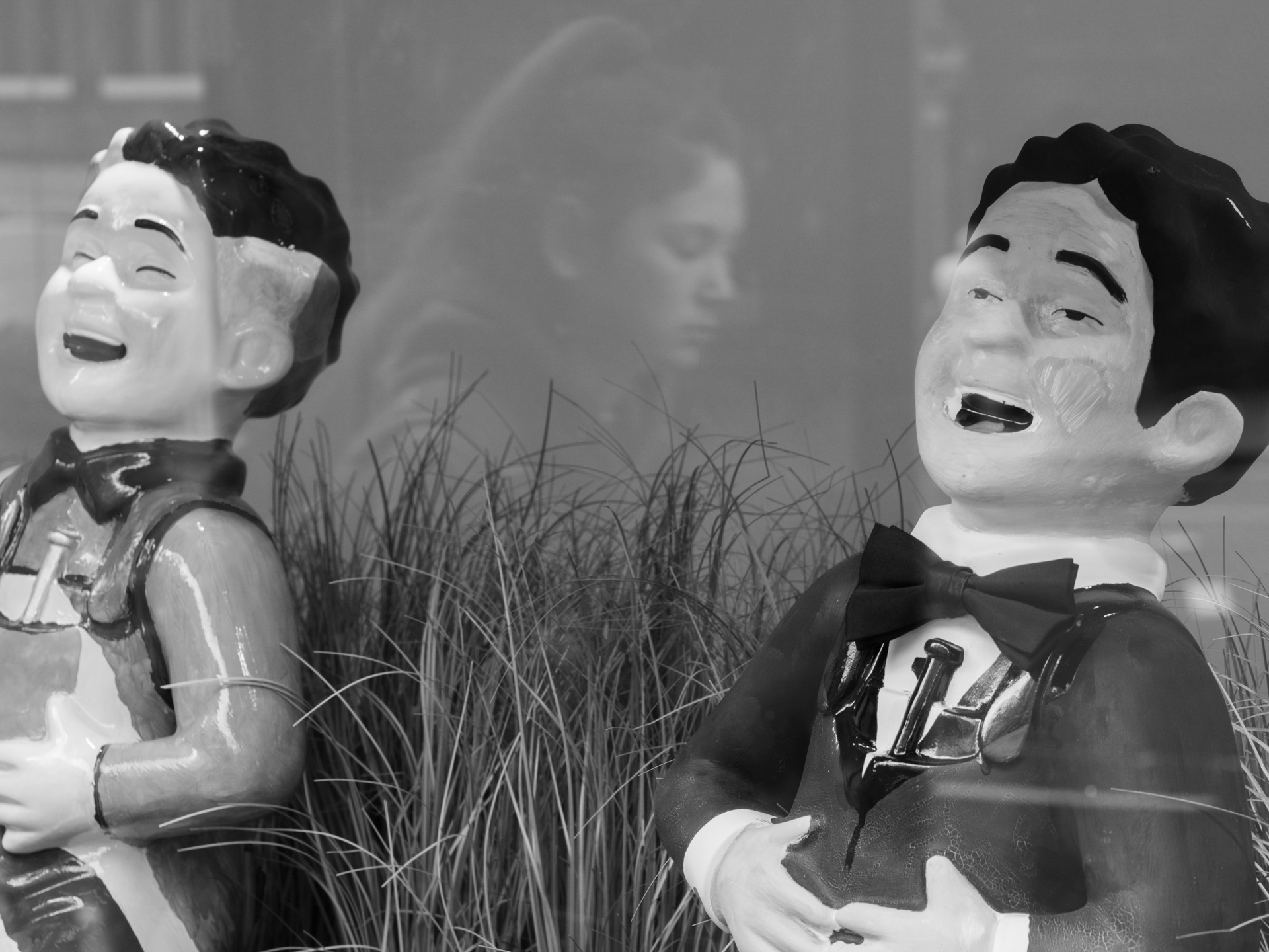 Two statuettes mock a passing girl reflected in a window.