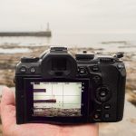 An OM-1 Mark II camera being held in the palm of a hand, looking at a pier and lighthouse.