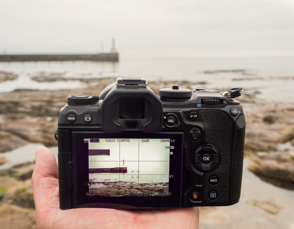 An OM-1 Mark II camera being held in the palm of a hand, looking at a pier and lighthouse.