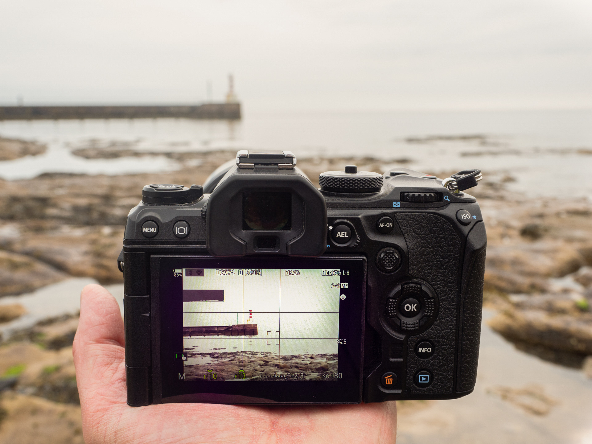 An OM-1 Mark II camera being held in the palm of a hand, looking at a pier and lighthouse.