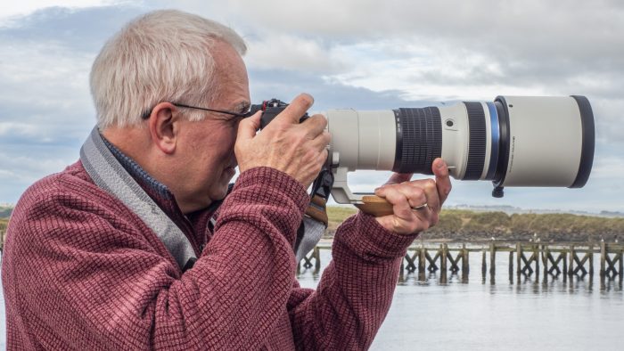 Ivor Rackham, photographer, stands with an OM-1 Mark II and the OM System 150-400 mm F4 PRO lens