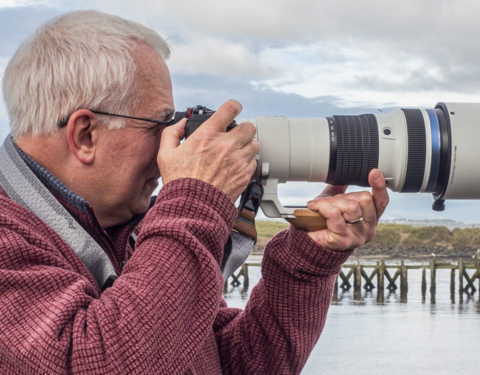 Ivor Rackham, photographer, stands with an OM-1 Mark II and the OM System 150-400 mm F4 PRO lens