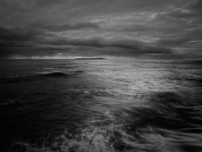 a moody black and white photo of the sea with reflected light leading the eye to Coquet Island in the distance