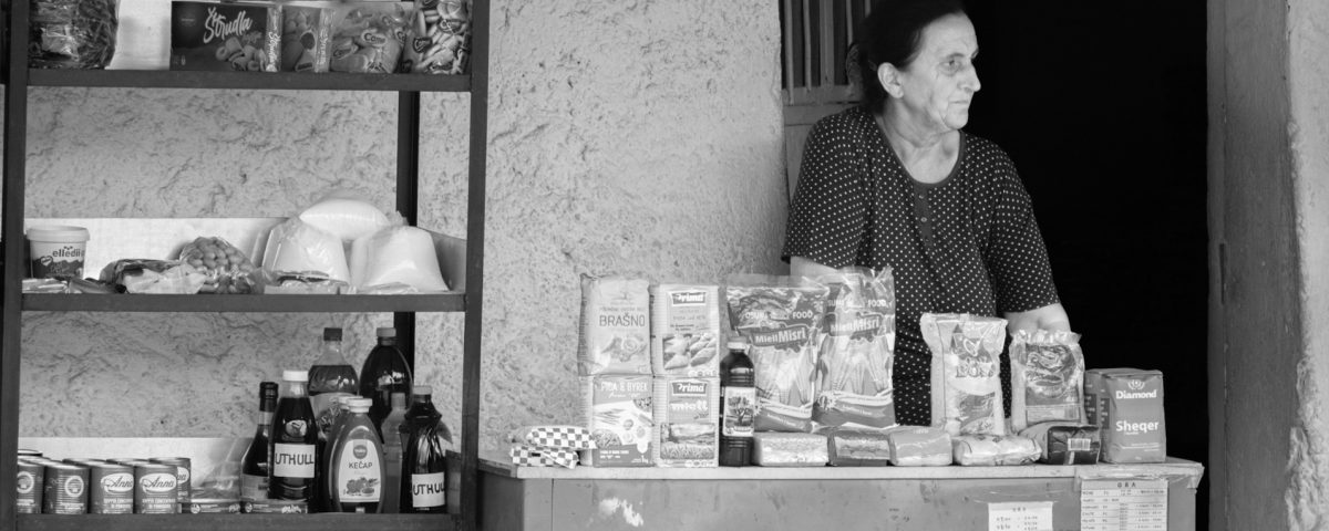 A woman street vendor stands behind her outdoor store selling groceries. She is staring at something to the right, out of the frame
