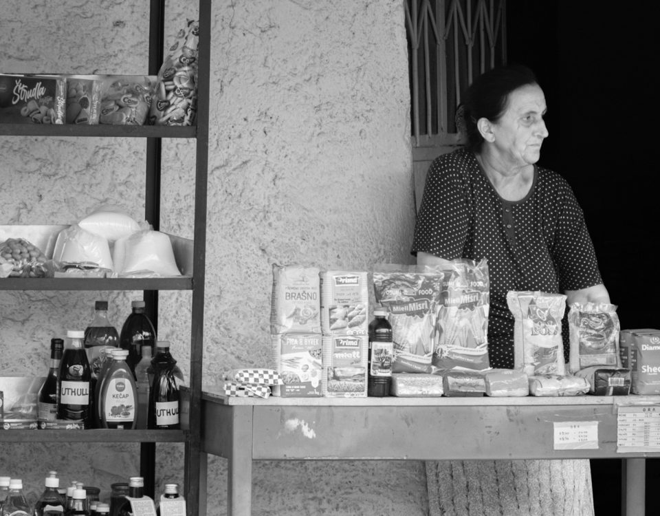 A woman street vendor stands behind her outdoor store selling groceries. She is staring at something to the right, out of the frame
