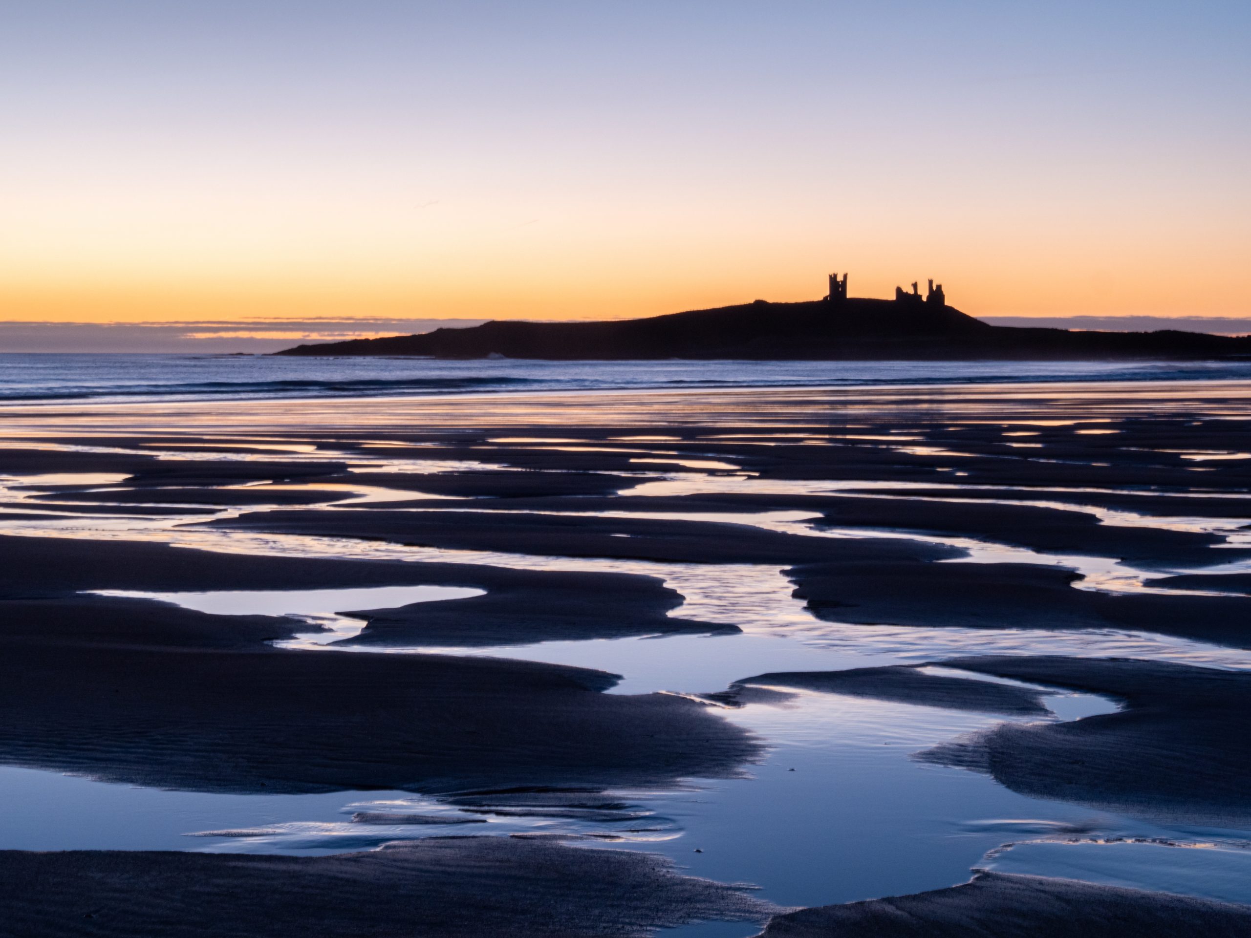 Dunstanburgh Castel at dawn from the beach with a network of water pools.