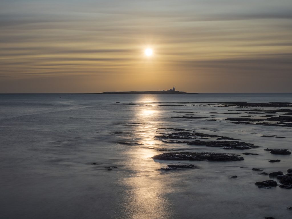 A long exposure image of the sun rising behind thin, smokey cloudss over the sea.