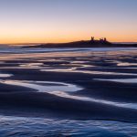 Patterns of water in the sand, on the beach at dawn. Dunstanburgh Castle sits on the headland on the horizon, with an orange glowing sun behind.