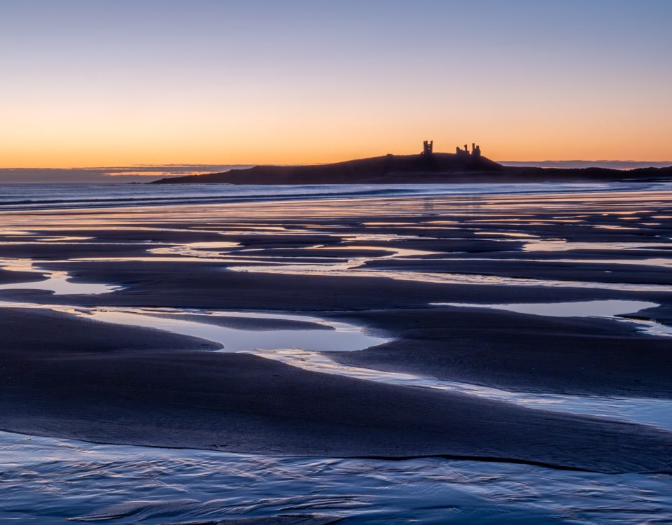 Patterns of water in the sand, on the beach at dawn. Dunstanburgh Castle sits on the headland on the horizon, with an orange glowing sun behind.