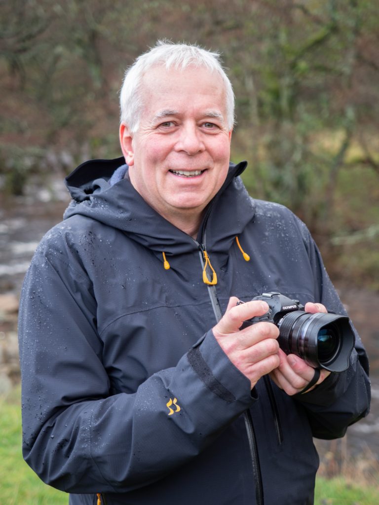 A portrait of Ivor Rackham in the rain, holding his OM System OM-1, which is IP53 weather sealed and so can withstand the downpour