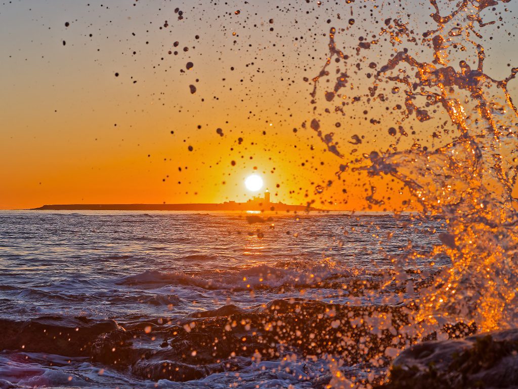 An HDR photo of a sunrise behind Coquet Island. A wave splashes in the forground. Shot with an OM-1 Mark II