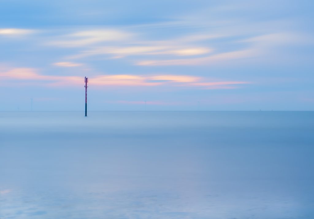 First Light, a soft, long exposure of the sunrise over the creamy smooth sea. A beacon port navigation post with a red top stands in the water, 