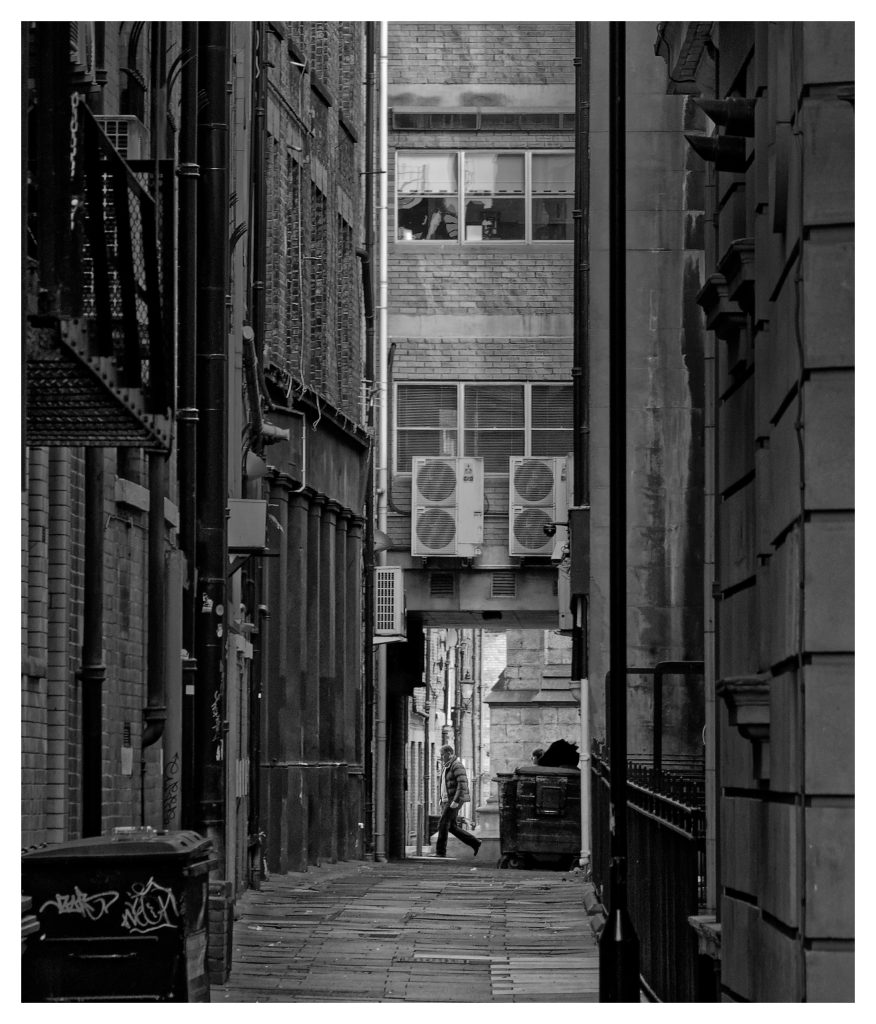 Photographed on a Street Photography class in Newcastle-upon-Tyne, a black and white image of a seedy-looking alleyway. a person in a puffer jacket crosses the end of the alley. Some classes in Ivor Rackham's Northumberland Photography Workshops venture further afield to Tyneside and Scotland.