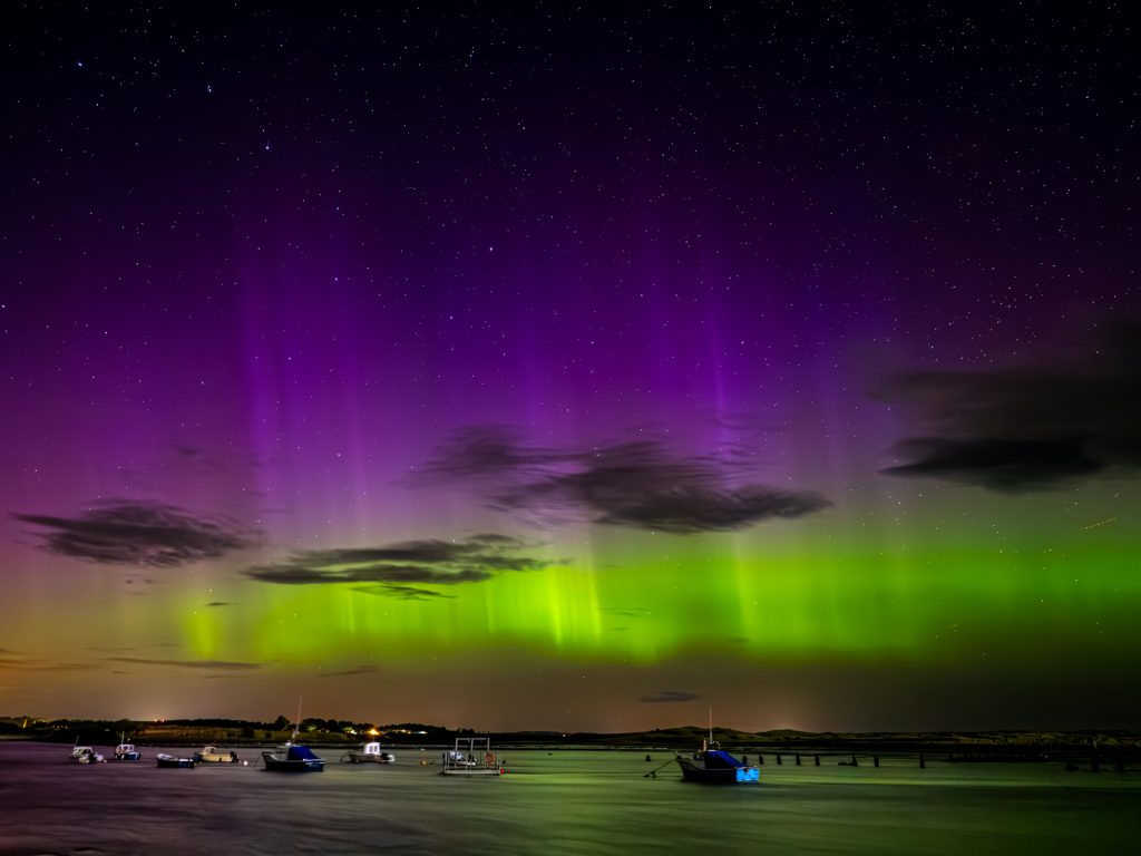 The Northern Lights with purple and green colours above water with boats moored. Above the aurora, stars are in the sky and Ursa Major (The Great Bear, called the Big Dipper in the USA) is clearly visible. Shot using an OM System camera