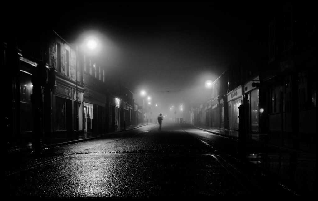 A night scene, a solo person walks up a dark and misty street. Typical of the type of photography on a Northumberland Photography Workshops
