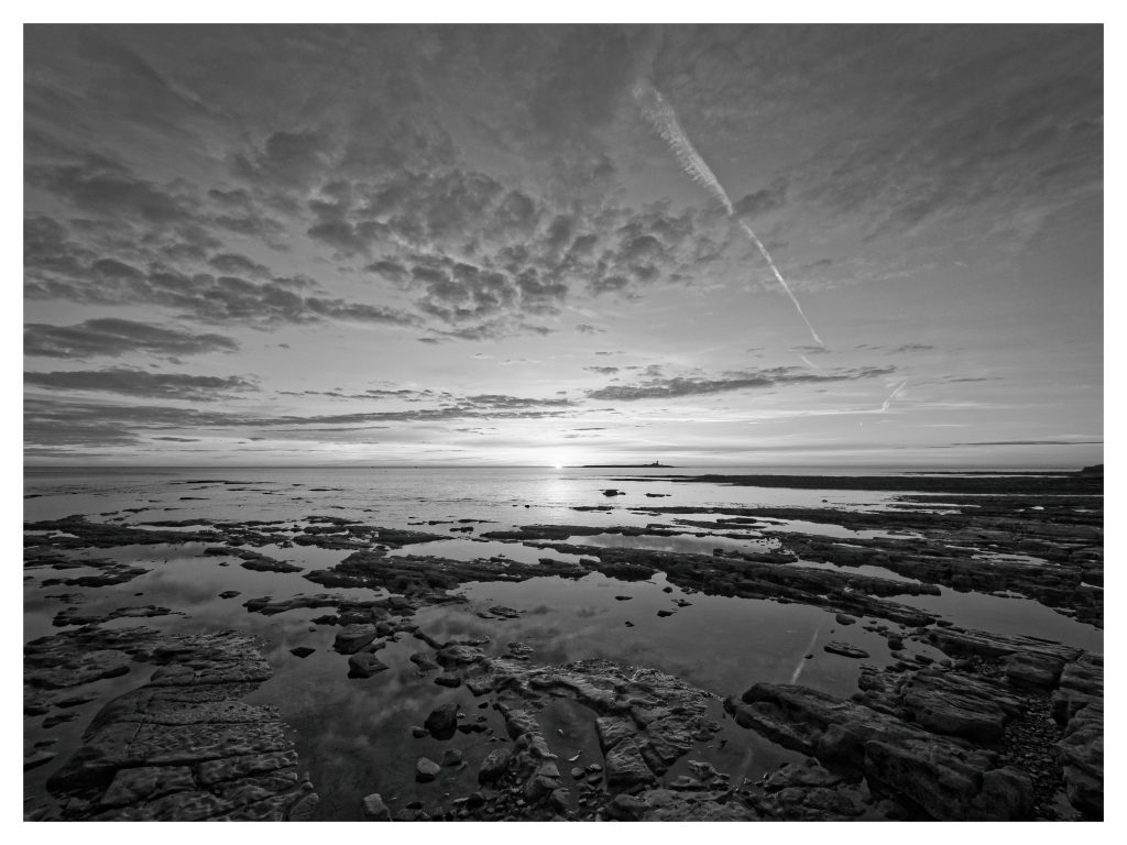 A black and white image of a beach at sunrise. Coquet Island is in the distance. Photographed with an OM System camera.