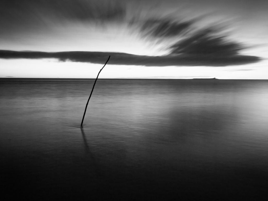 Northumberland Photography Workshops - A surreal black and white long exposure image of a long exposure showing a stick in the sea water. In the distance an island sits on the horizon. Clouds are stretched across the sky during the 60 seconds that the shutter was open.