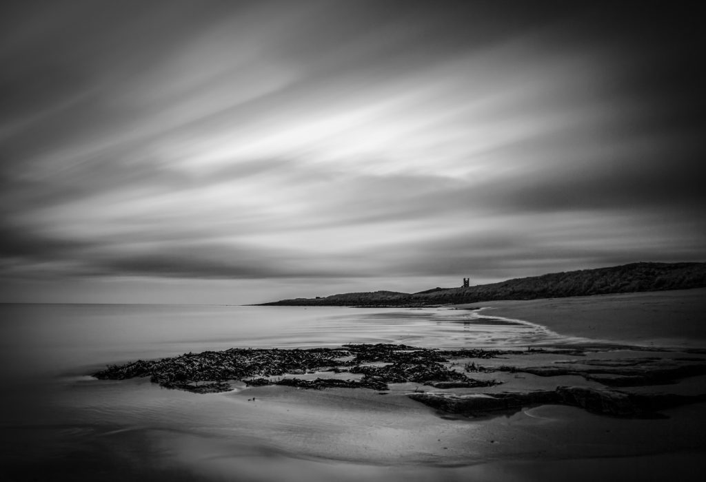 A long exposure black and white photo of a beach. The clouds are elongated. A solitary tower sits on a headland in the distance. Shot with an OM System Camera,  OM-1 Mark II using Live Time