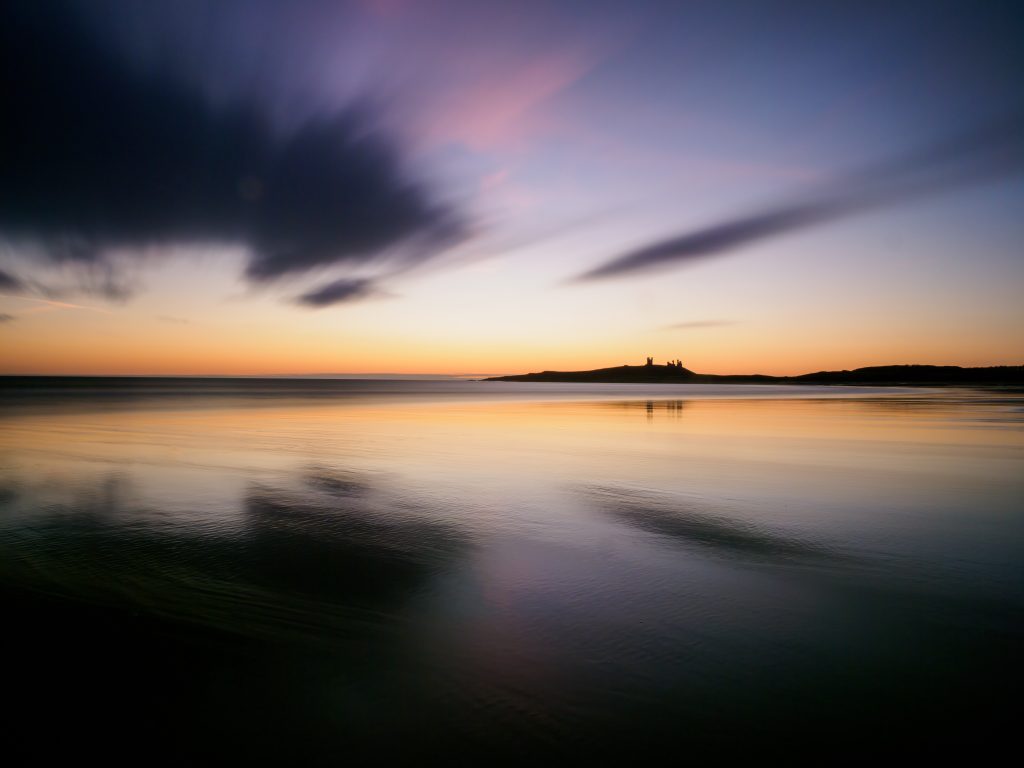 A long exposure of a beach, at dawn showing moving clouds. In the distance, a castle sit of a headland.