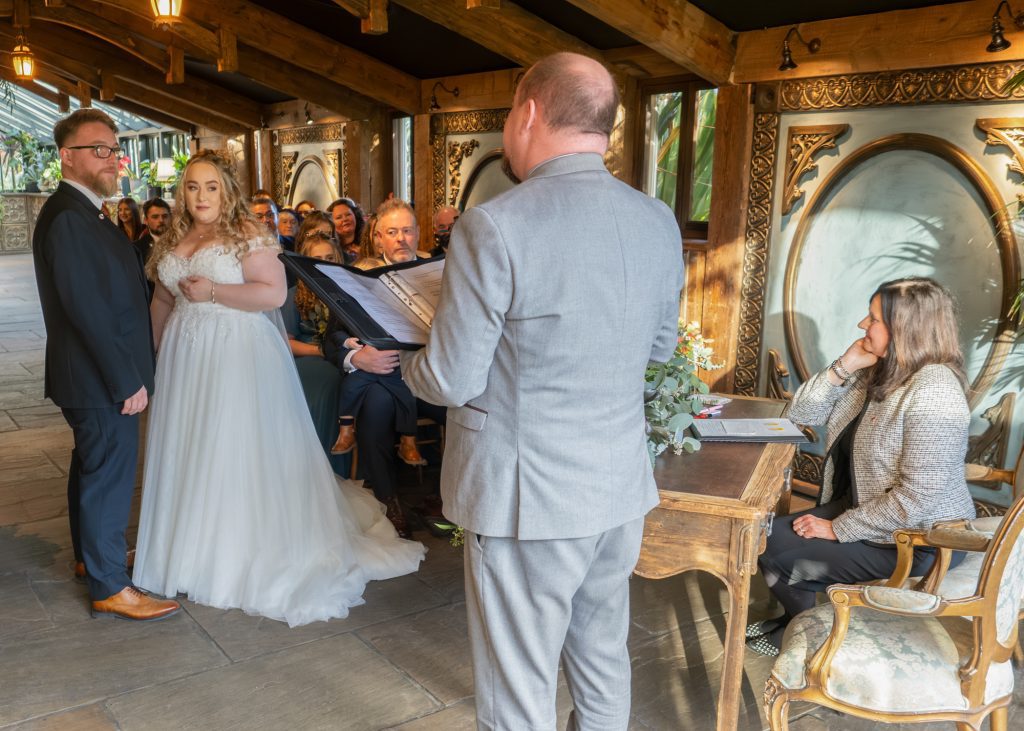 A wedding scene with the bride and groom listening to the man running the ceremony. The congregation watches on. Shot in low light with a high ISO using an OM System camera which copes exceptionally well with low light,
