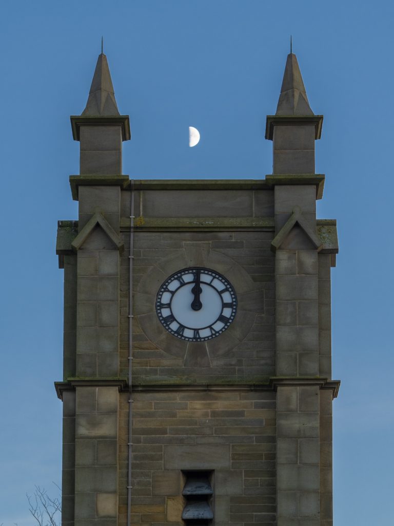 A clock tower shows midnight. the hands point to a half moon above it in the night sky. Shot with an OM System camera