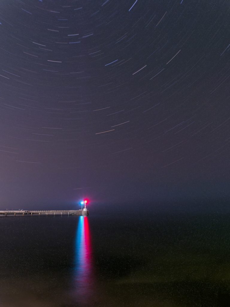 A long exposure of beacons at the end of two piers on a misty evening. Star trails circle around in the sky. 